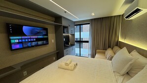 Ein modernes Hotelzimmer mit einem großen Bett, einem flachbildfernseher an der Wand, einer kleinen Küchenzeile und Blick durch ein großes Fenster.