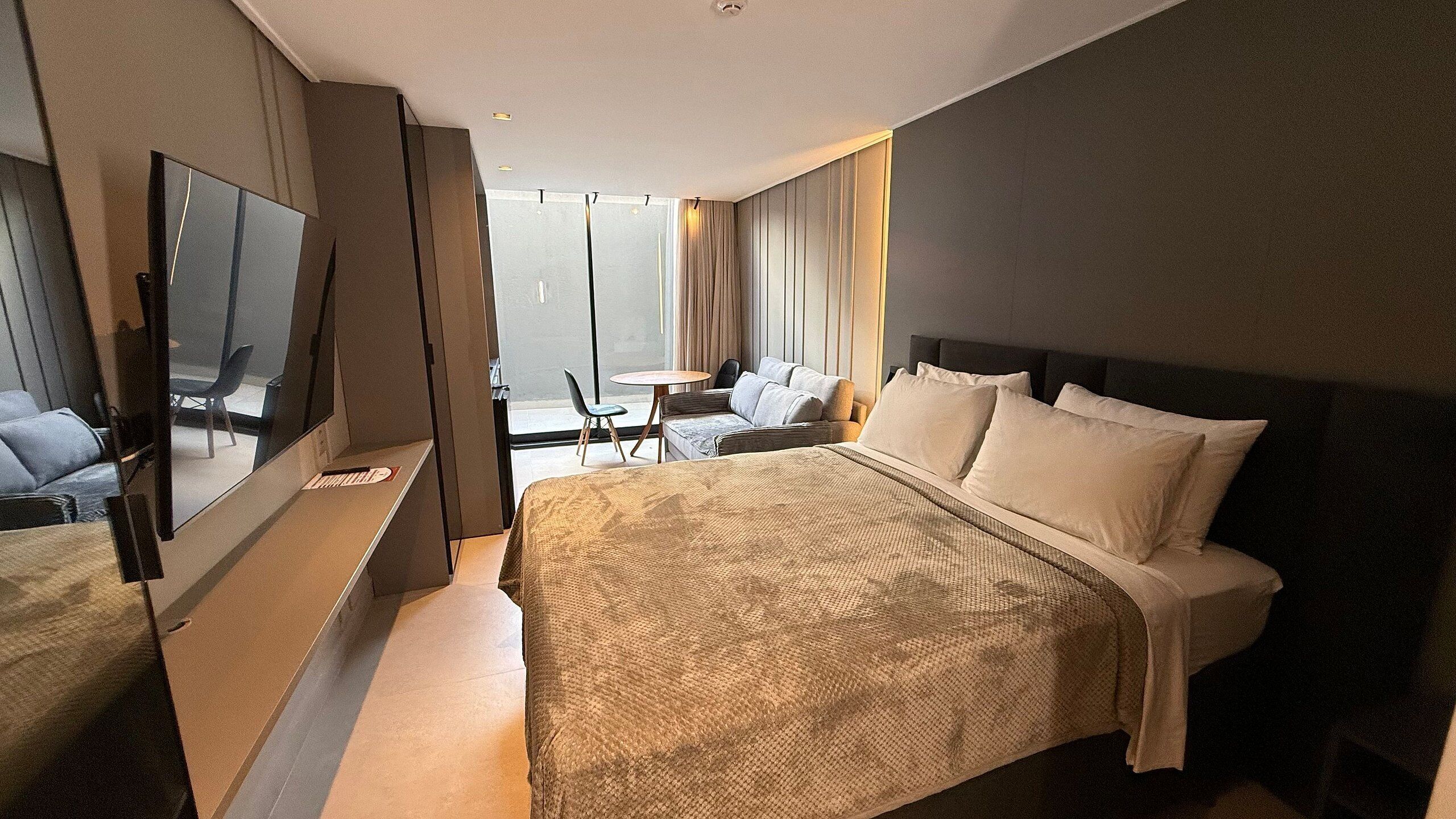 A hotel room with a large bed, a TV, a seating area with a table and chairs, and a view through a glass door.