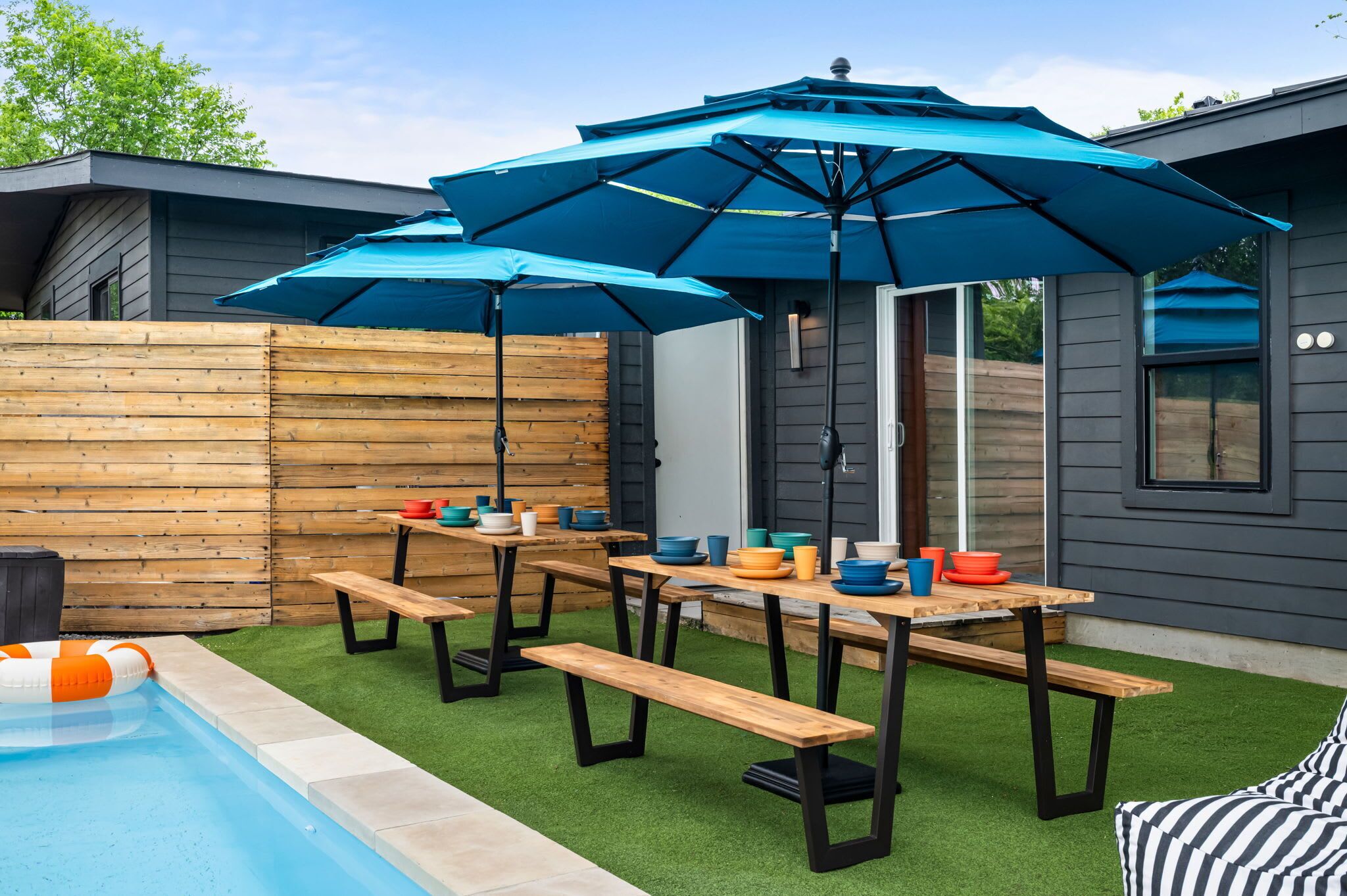 A backyard with a pool, wooden deck, picnic tables, and blue umbrellas.