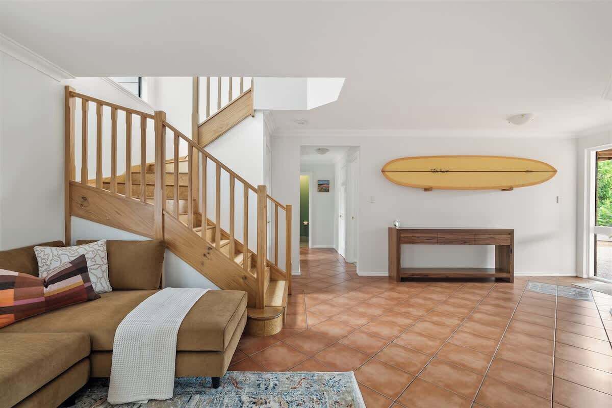 A staircase leading to an upper floor, a wooden bench, a surfboard on the wall, and a brown sofa with cushions.
