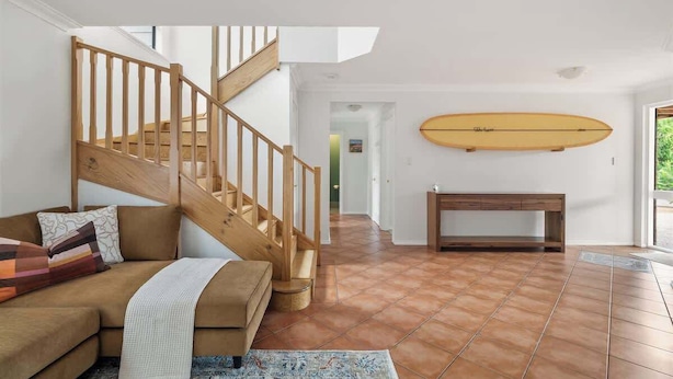 A staircase leading to an upper floor, a wooden bench, a surfboard on the wall, and a brown sofa with cushions. - Canopy House—Refined Bushland Haven near the Coast (Quedjinup)