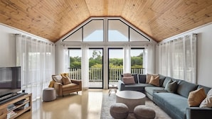 A modern living room with a sloped ceiling, a large TV, a sofa, a coffee table, and a balcony with a view. - Canopy House—Refined Bushland Haven near the Coast (Quedjinup)