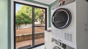 A laundry room with a front-loading washer and dryer, a sliding glass door leading to a patio with a brick wall and greenery. - Canopy House—Refined Bushland Haven near the Coast (Quedjinup)
