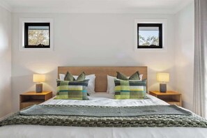 Un dormitorio con cabecera de madera, dos mesitas de noche con lámparas, una cama con manta verde y dos ventanas pequeñas con vistas a árboles.