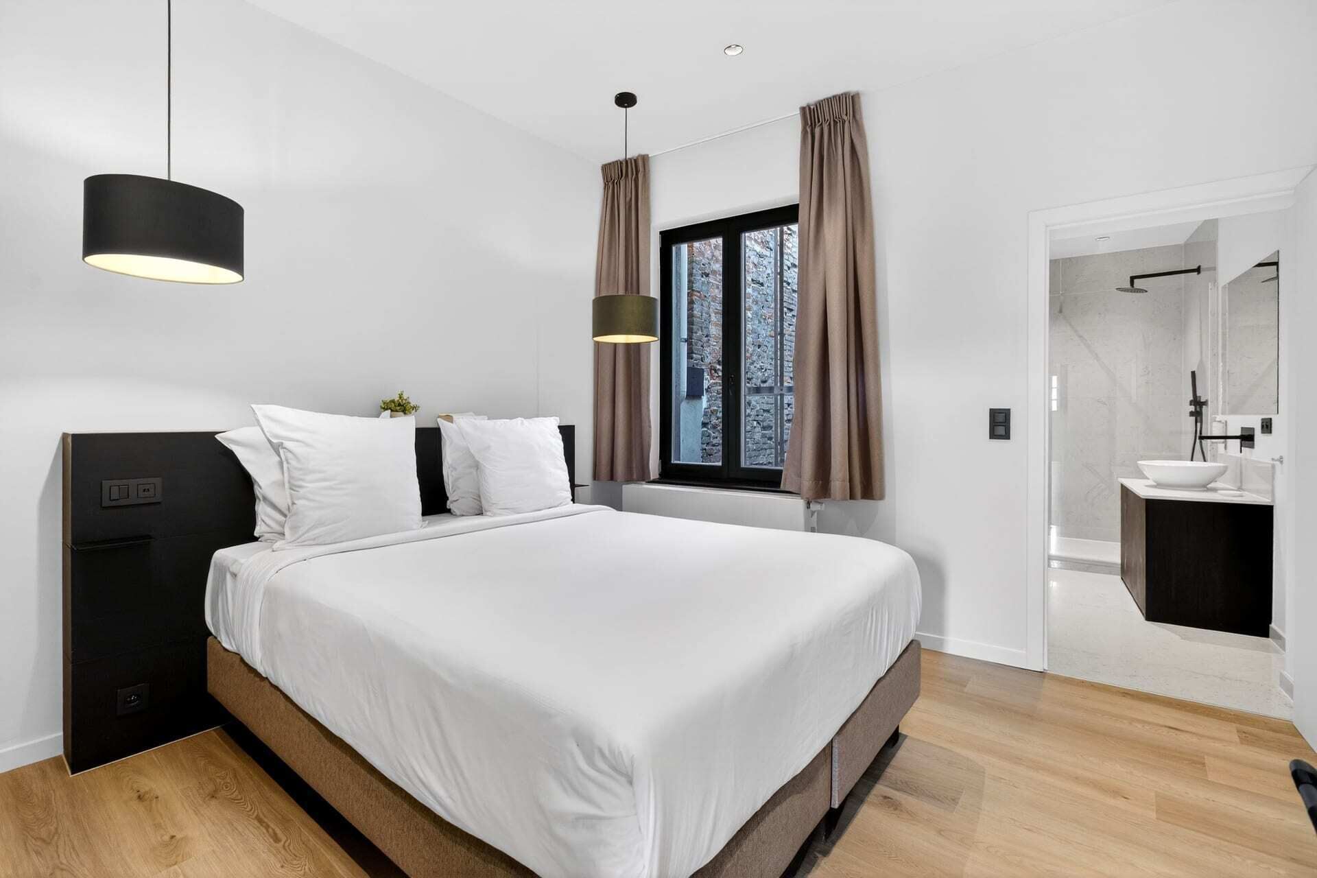 A modern bedroom with a large bed, a bedside table, a hanging light fixture, a window with curtains, and a bathroom with a sink and mirror.