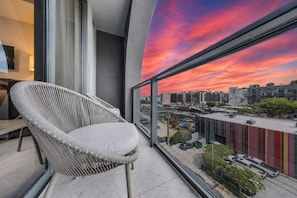 A balcony with a wicker chair, a cityscape view, and a colorful sunset. - Modern Wynwood 1BR | Rooftop Pool & Gym (Miami)
