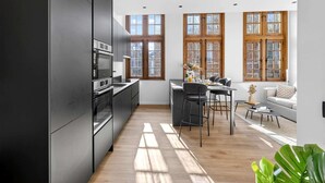 A modern kitchen with dark cabinetry, a dining area with a table and chairs, and large windows with wooden frames. - Maison Grand-Place 2 (Brussels)