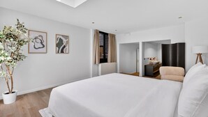 Ett modernt hotellrum med en stor säng, en krukväxt, abstrakt konst, ett badrum med handfat och spegel samt utsikt genom en öppen dörr.