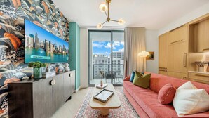 Een moderne woonkamer met een bank, een salontafel, een televisie op een kast, en uitzicht op de stad door een groot raam.