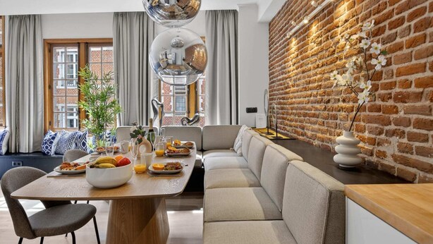 Un salon moderne avec un mur en briques, un canapé, une table à manger où est servi le petit-déjeuner, et une grande fenêtre.