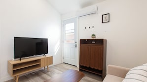 A modern living room with a flat-screen TV on a wooden stand, a wooden table, a beige sofa, a wardrobe, and a door with a window.