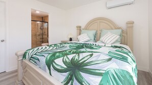 A bedroom with a bed, a nightstand, a lamp, and a bathroom visible through an open door.