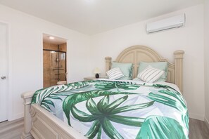 Un dormitorio con cama, mesita de noche, lámpara y baño visible a través de una puerta abierta.