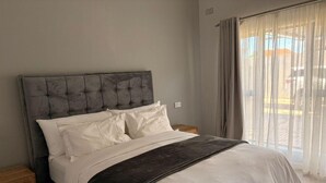 A bedroom with a tufted headboard, a bed with white bedding, a dark throw blanket, wooden nightstands, and a view through sheer curtains. - One Bedroom Modern Apartment in Fomona - 2386 (Bulawayo)