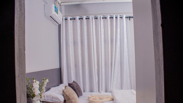 Uma cama pequena e bem arrumada, com almofadas e um cobertor, um vaso com flores e uma cortina.
