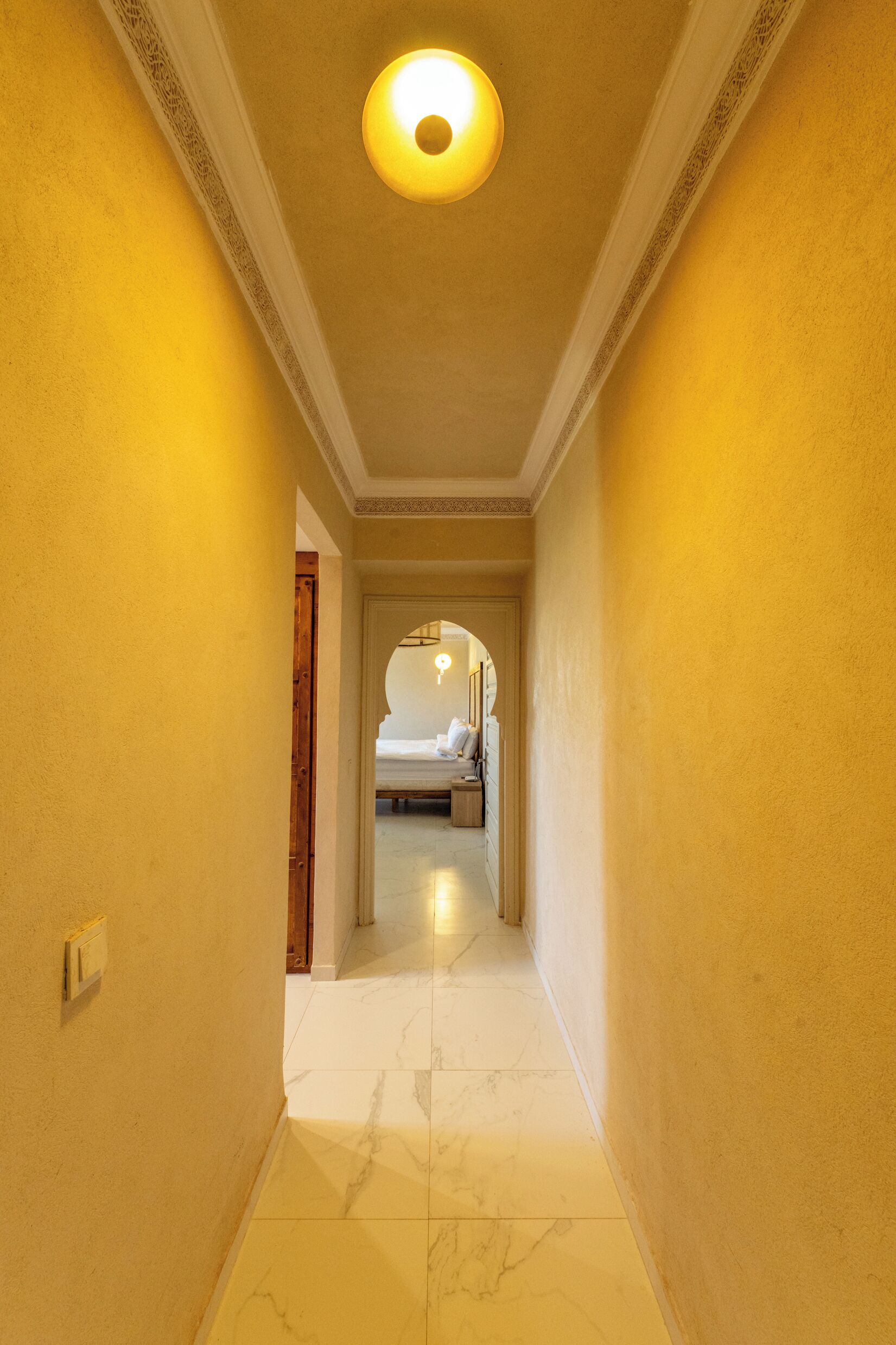 Un couloir au sol en marbre qui mène à une pièce contenant un lit et une table de chevet.