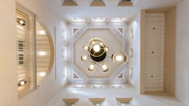 A room with a geometric patterned ceiling, a central chandelier, and a window with decorative blinds. - Villa Aurea (Marrakech)