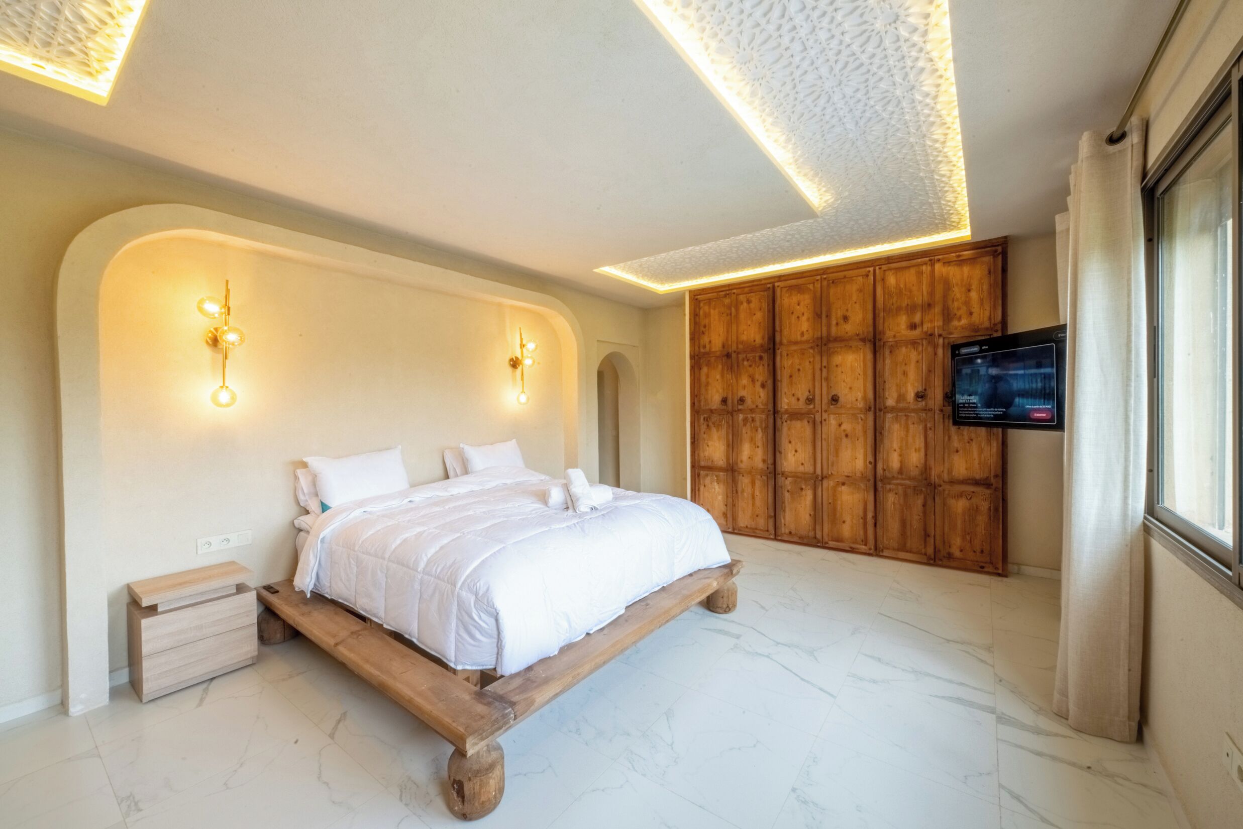 A bedroom with a bed, wooden headboard, wall-mounted lights, a television, and a window with curtains.