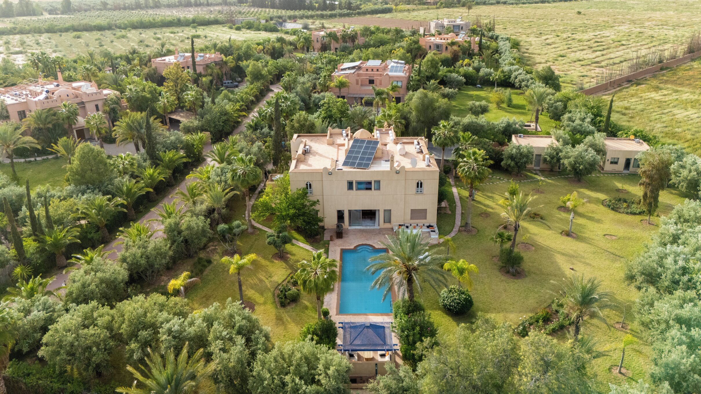 An aerial view of a residential area with multiple houses, a central building with a swimming pool, and surrounding greenery.