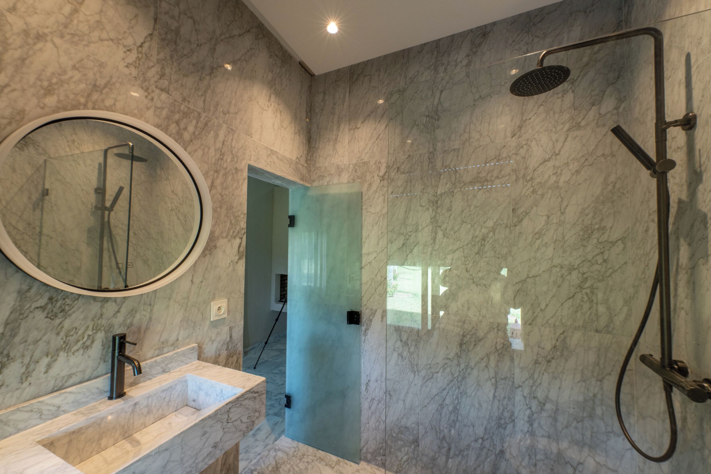 A bathroom with a marble wall, a round mirror, a sink, and a shower.