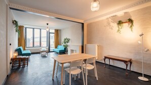 A modern room with a wooden table, white chairs, a blue sofa, and a small table with a lamp. - 2BR City Stay - Grand Central Area (Antwerp)