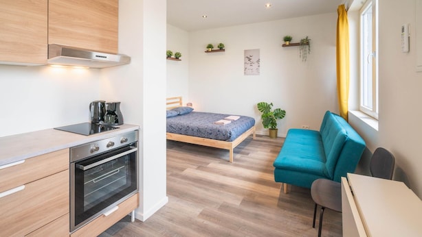 Ein kleines Zimmer mit einer Küchenzeile, einem Bett, einem Schlafsofa und einem Fenster mit gelben Vorhängen.