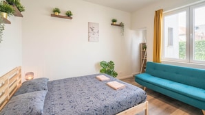 Ein Schlafzimmer mit einem Bett, einem blauen Sofa, einem Fenster mit Vorhängen und an der Wand befestigten Regalen mit Pflanzen.