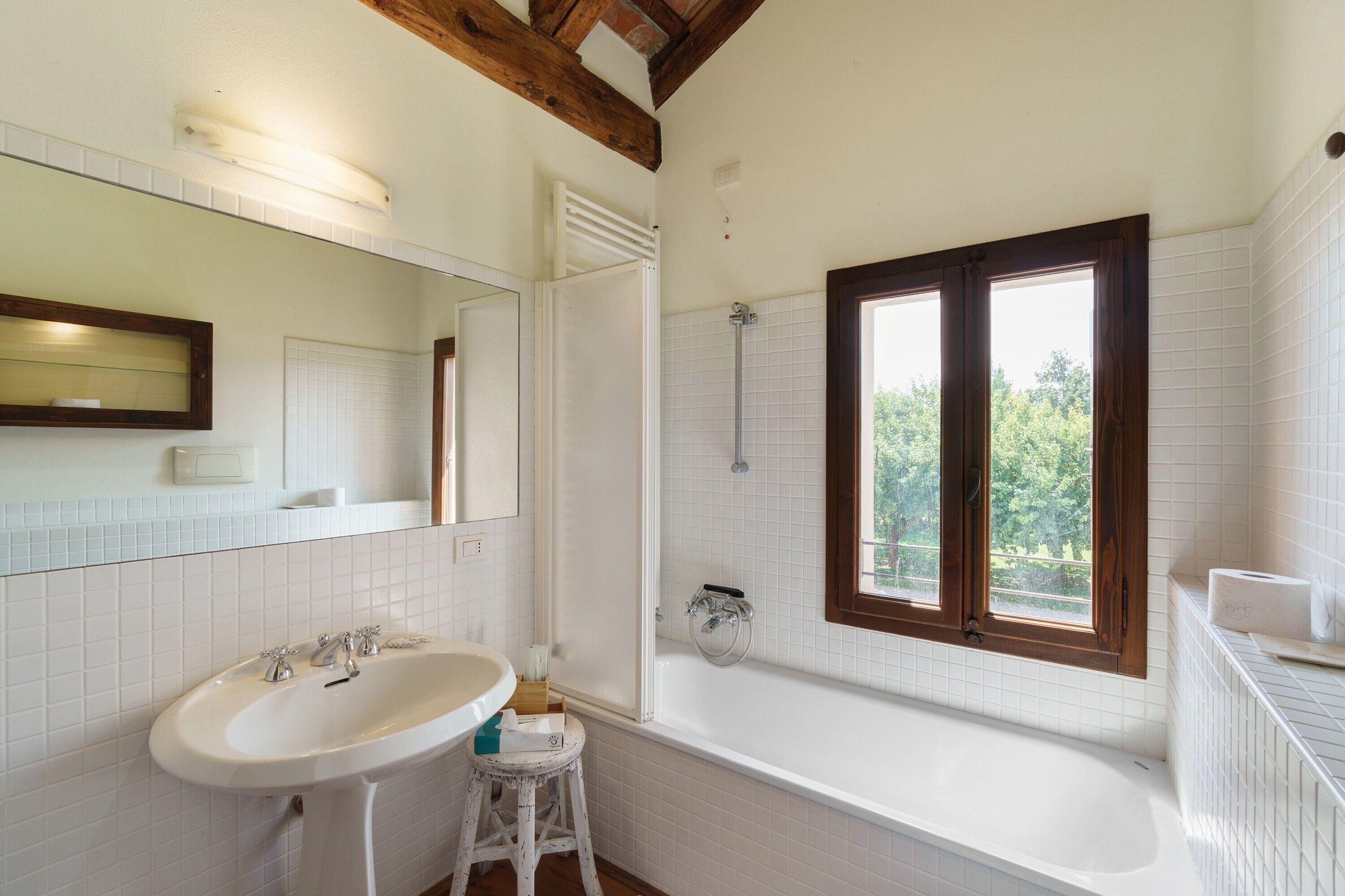 Un baño con un lavabo blanco, una bañera, un ventanal con vistas a la vegetación y una viga de techo de madera.