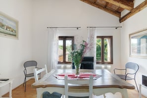 A dining room with a wooden table, white chairs, and a vase of flowers. There are two windows with curtains, a fireplace, and a painting on the wall. - 'Grimilda' Apartment with Shared Pool, Wi-Fi and Air Conditioning (Piombino Dese)