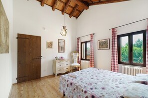 A bedroom with a wooden door, a dresser, a chair, a bed with floral bedding, a window with curtains, and a radiator. - 'Grimilda' Apartment with Shared Pool, Wi-Fi and Air Conditioning (Piombino Dese)