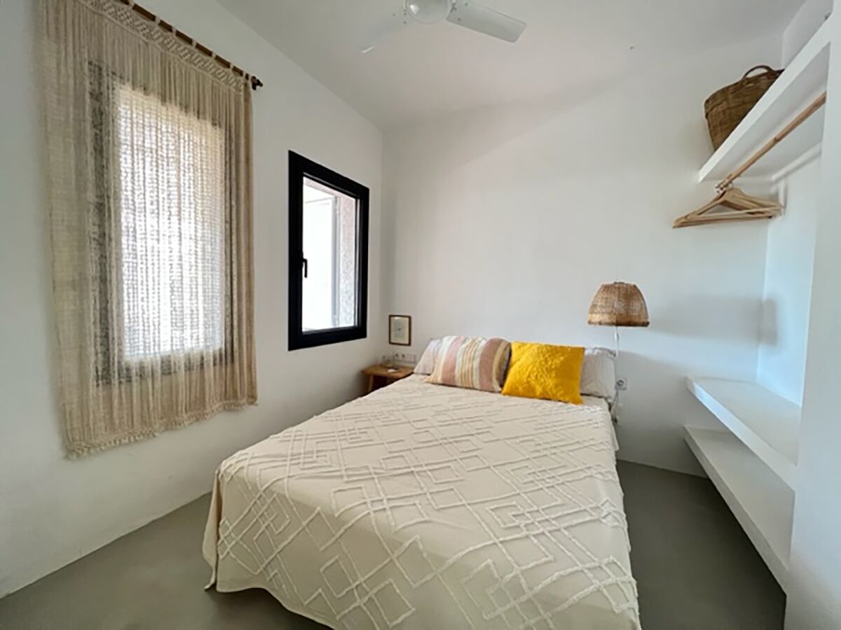 A bedroom with a bed, a window with curtains, a bedside table, and a shelf with a basket.