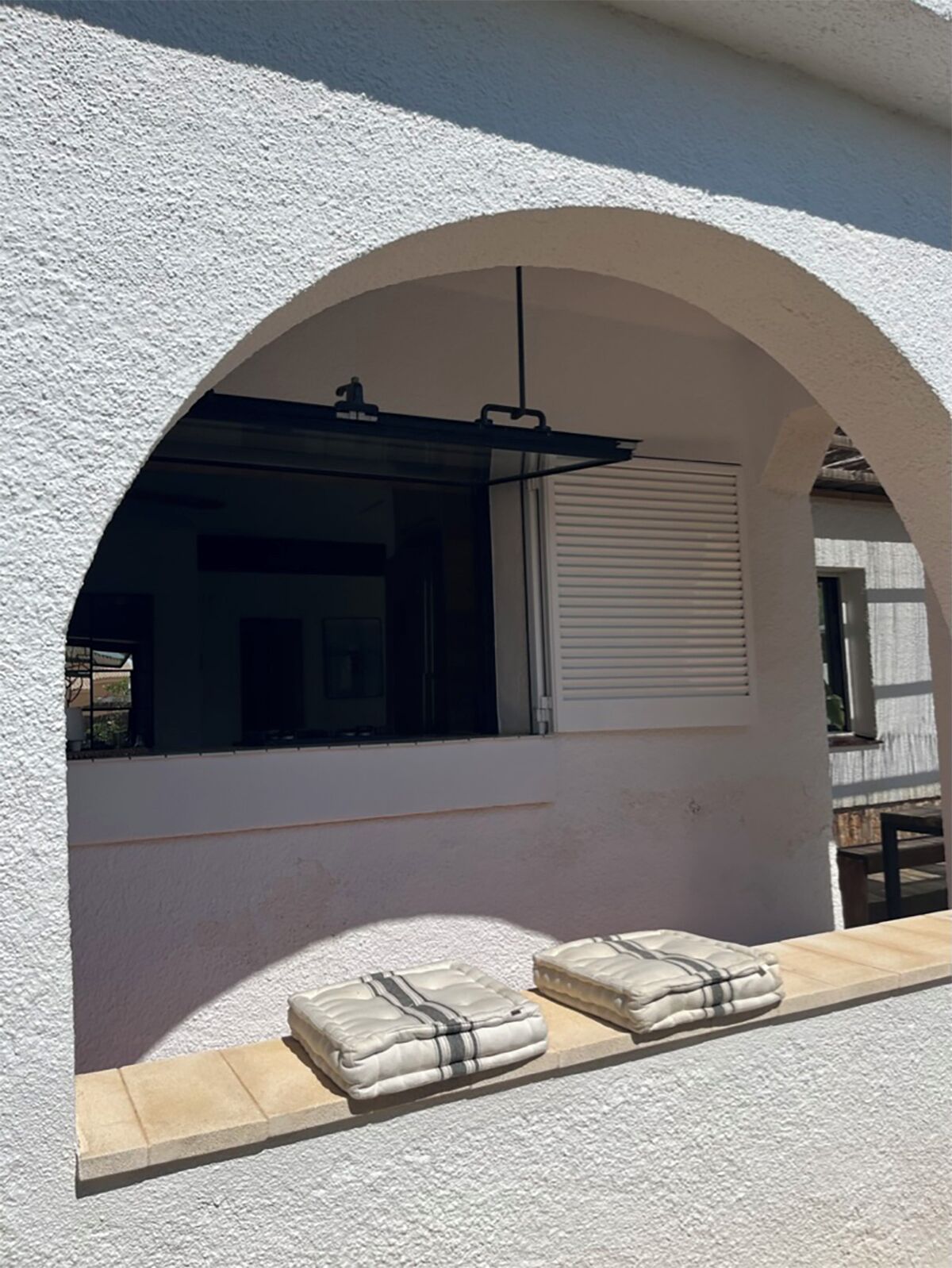Una ventana arqueada con vista a una habitación, un persiano cerrado y dos colchones sobre un alféizar.