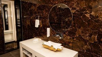 Ein Badezimmer mit Marmorwand, einem weißen Waschtisch, einer goldenen Spüle, einem runden Spiegel und einem Handtuchhalter.