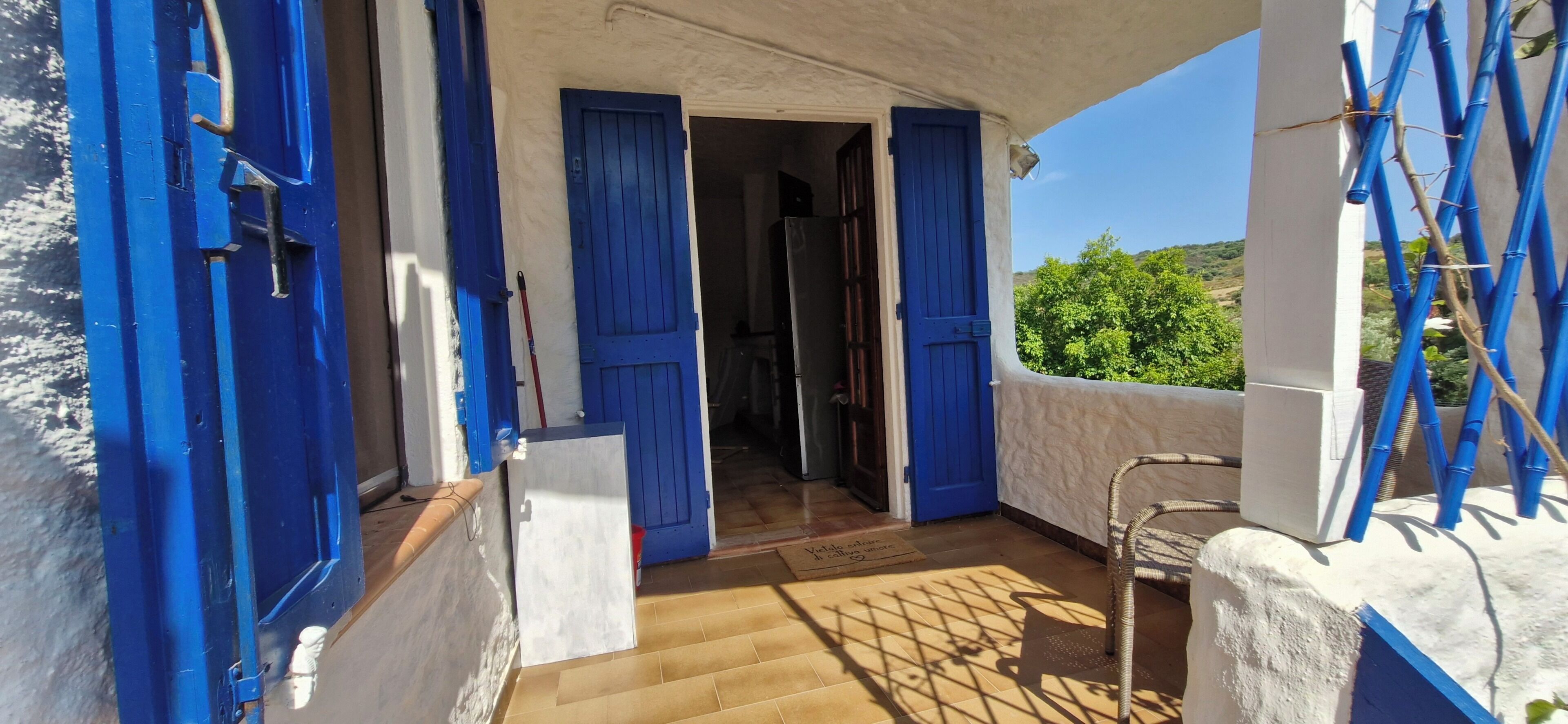 Un balcone con persiane blu, una sedia in vimini e vista sul verde.