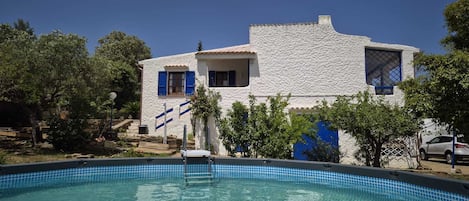A white building with blue shutters, a swimming pool, and surrounding trees.
