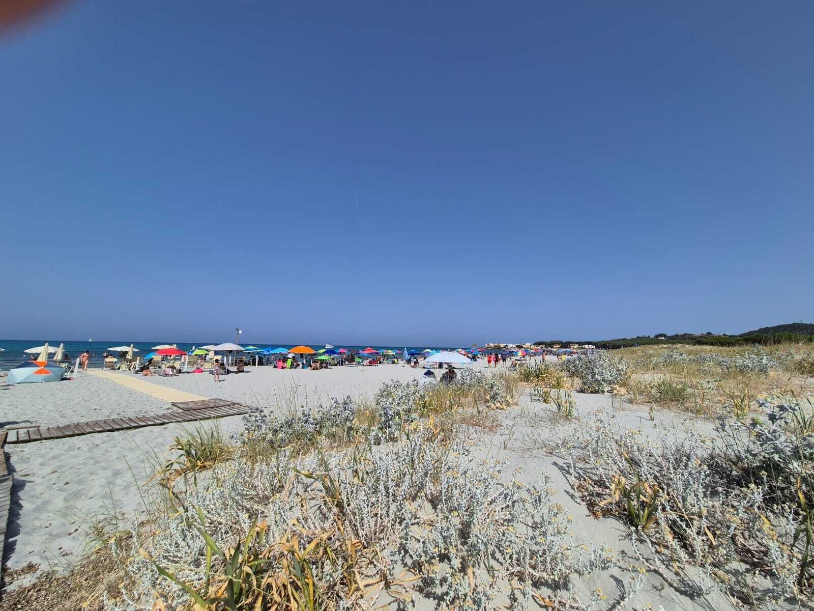 Una playa con sombrillas y gente, un sendero arenoso y vegetación escasa.