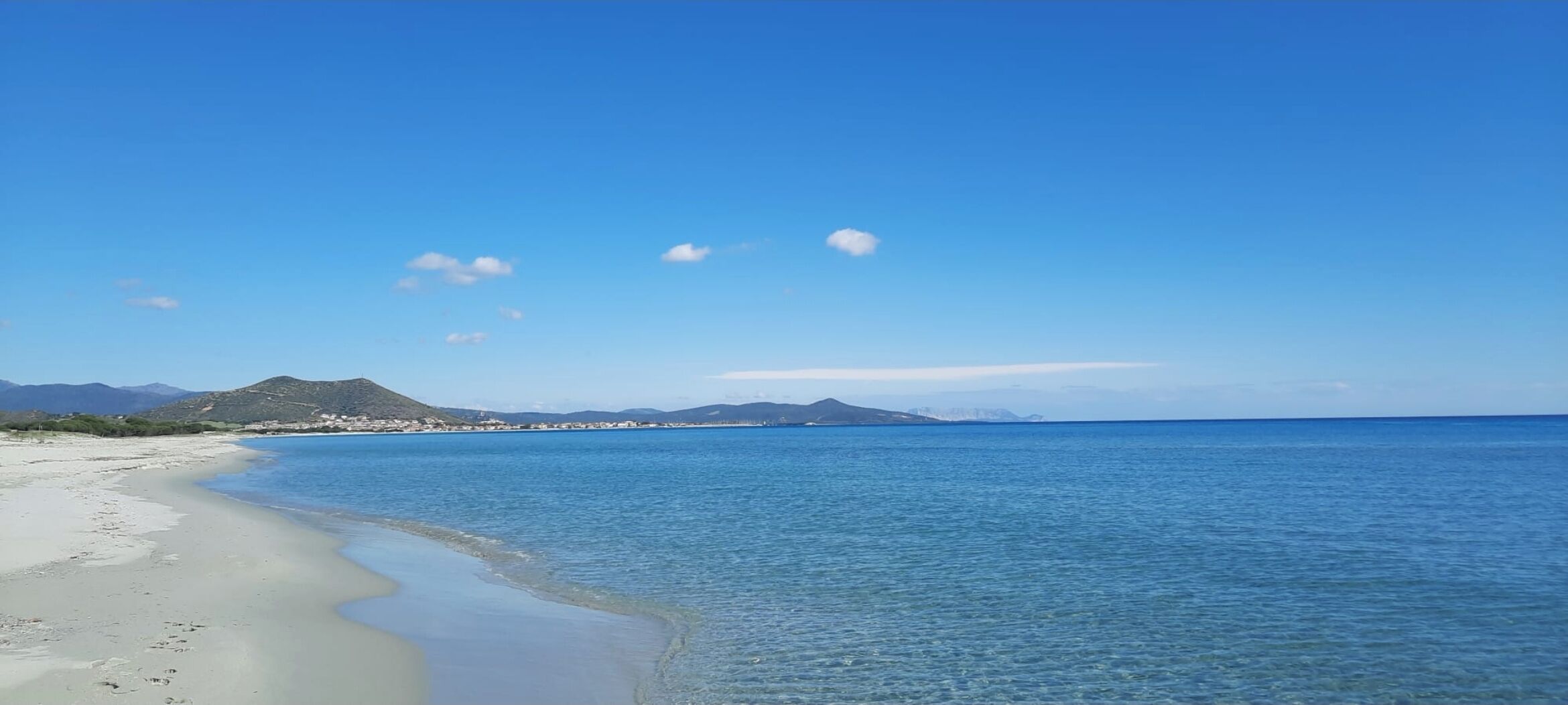 A beach with clear blue water, a sandy shore, and distant mountains under a clear sky.