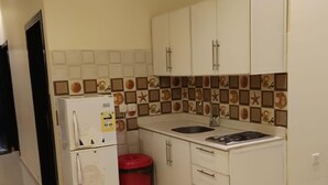 A compact kitchen with white cabinets, a sink, a refrigerator, and a red trash bin. - Quiet Rooms 2   (Riyadh)