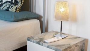 A bedside table with a lamp, a remote control, and a patterned cushion on a bed.