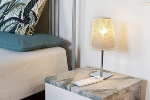A bedside table with a lamp, a remote control, and a patterned cushion on a bed.