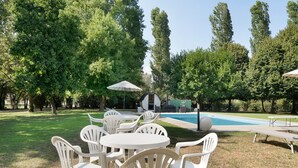 An outdoor seating area with white plastic chairs and tables, a pool, and a building with a green facade in the background.