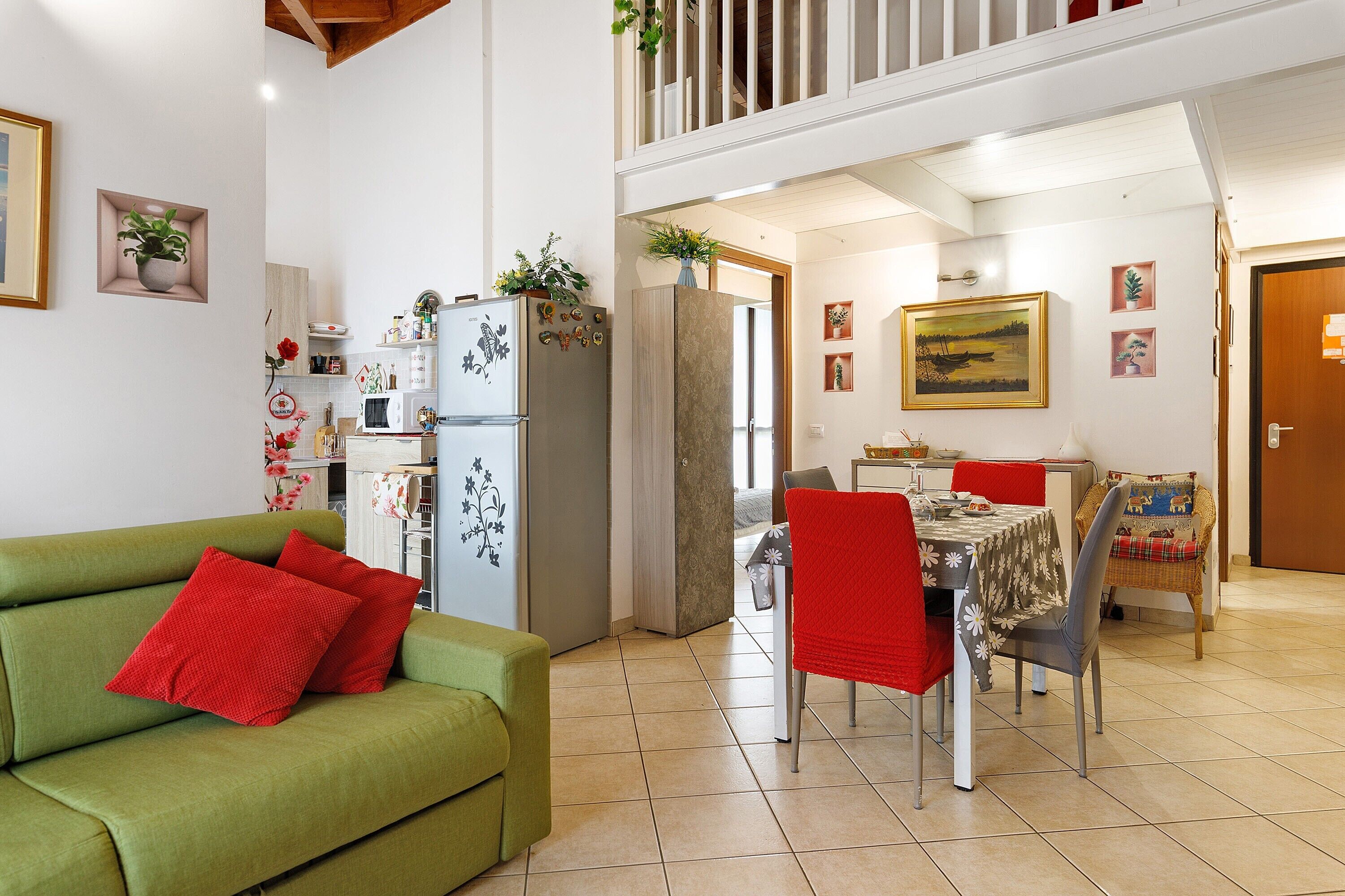 Un salon avec un canapé vert, des coussins rouges, un coin repas avec une table et des chaises, une cuisine équipée d’un réfrigérateur et un escalier menant à un étage supérieur.