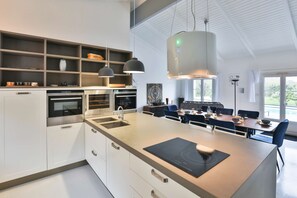 A modern kitchen with built-in appliances, a central island, pendant lighting, and a dining area with blue chairs.