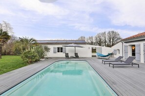 A swimming pool with lounge chairs and a hammock, surrounded by a wooden deck and a white building with shutters.