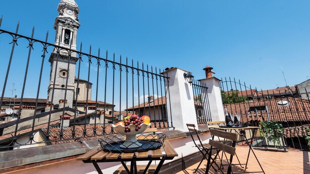 A terrace with a table set for dining, a decorative bowl of fruit, and a view of a historic clock tower. - Central Budget Rooms (Bergamo)