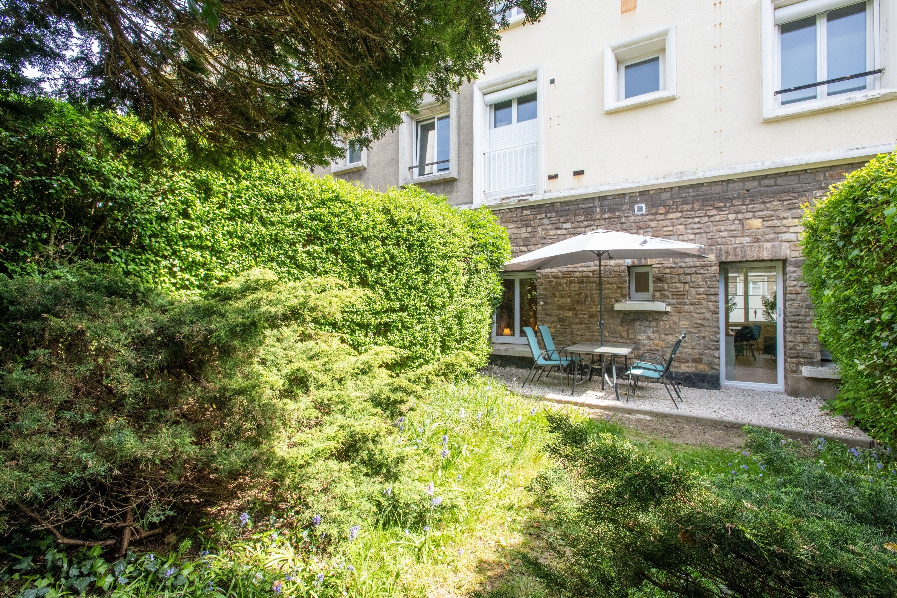 Une cour avec une table et des chaises sous un parasol, entourée d’une végétation luxuriante et d’un bâtiment en pierre.