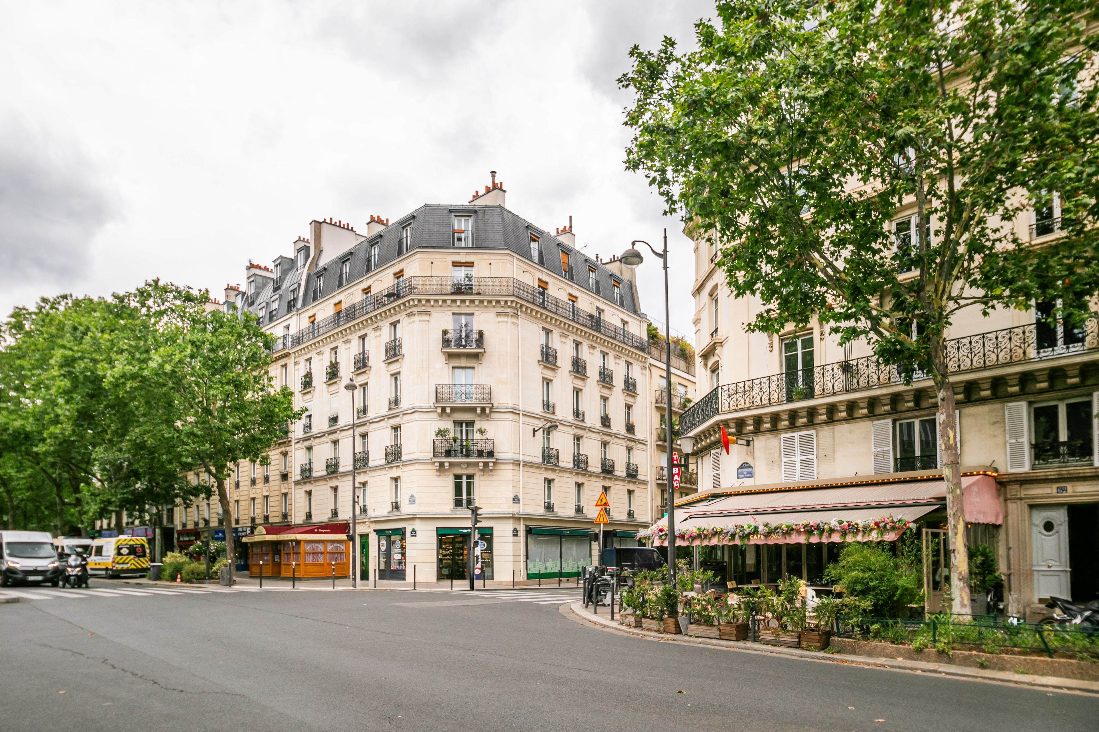 Vista de una calle con un edificio de varios pisos, balcones y una cafetería con mesas al aire libre.