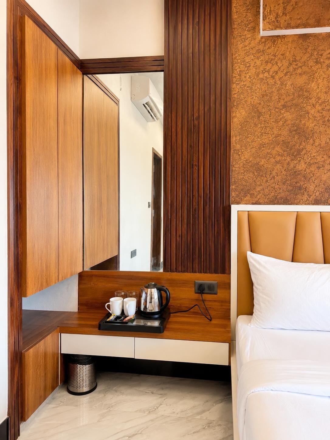 Habitación de hotel con una cama, un escritorio de madera con una bandeja de tazas y una tetera, un cubo de basura y un espejo grande.