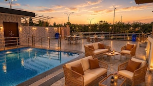 A pool area with wicker furniture and a patterned floor, surrounded by a stone wall and a view of the sunset.