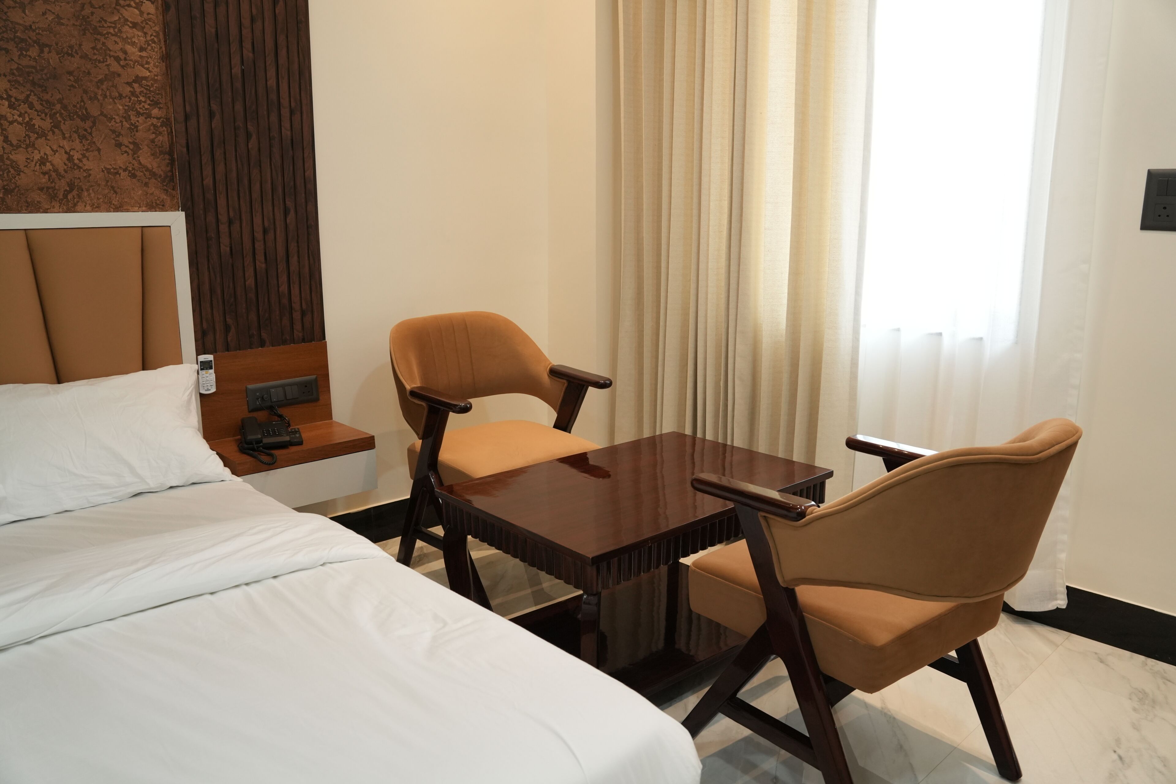 Quarto de hotel com uma cama, duas cadeiras, uma mesinha, um criado-mudo e uma janela com cortinas.